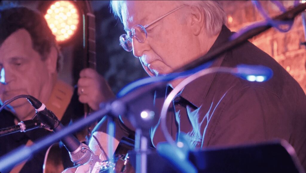 An elderly man playing guitar on stage with intense focus.